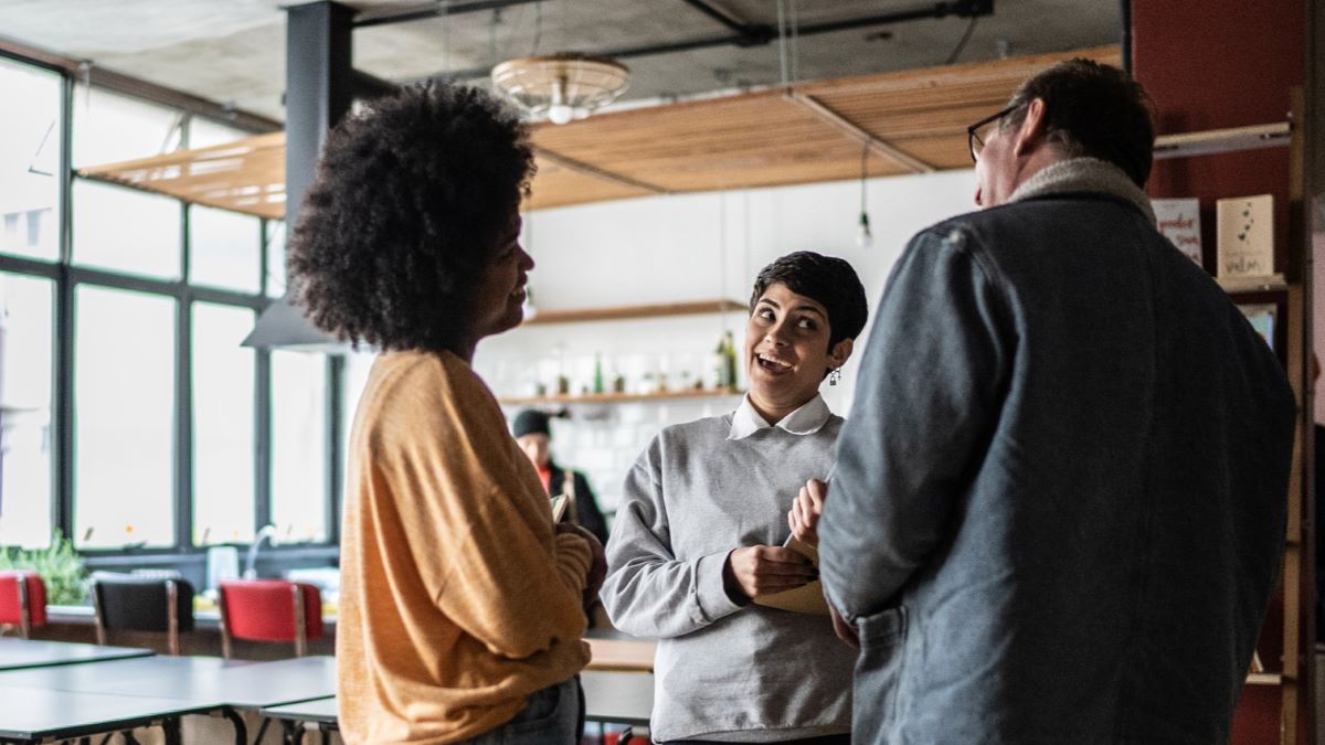 three people stand talking animatedly in a room with large windows