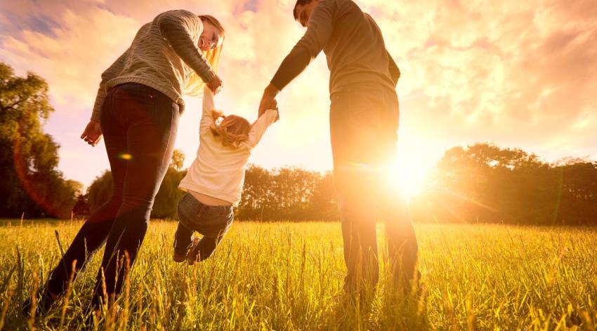 Two parents hold their child's hands and lift her up in a field as the sun sets.